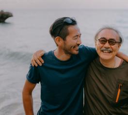 younger man standing next to an older man with his arm around his shoulder at the beach