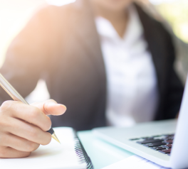 Closeup of laptop and pen writing in notebook with a person in the background